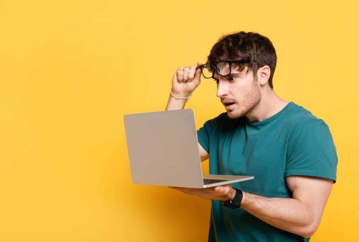 Man lifting glasses in surprise while looking at laptop