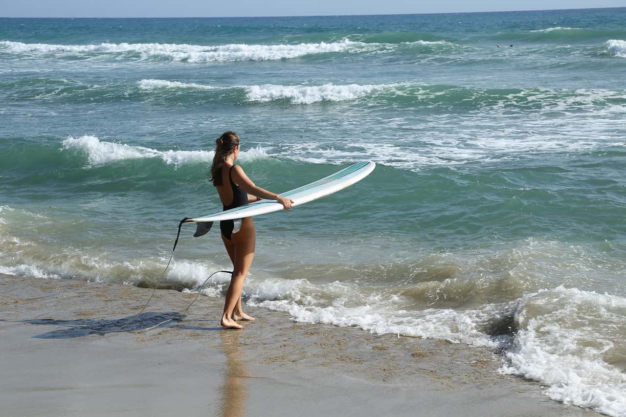 A woman holding a surfboard stands on the beach facing the waves