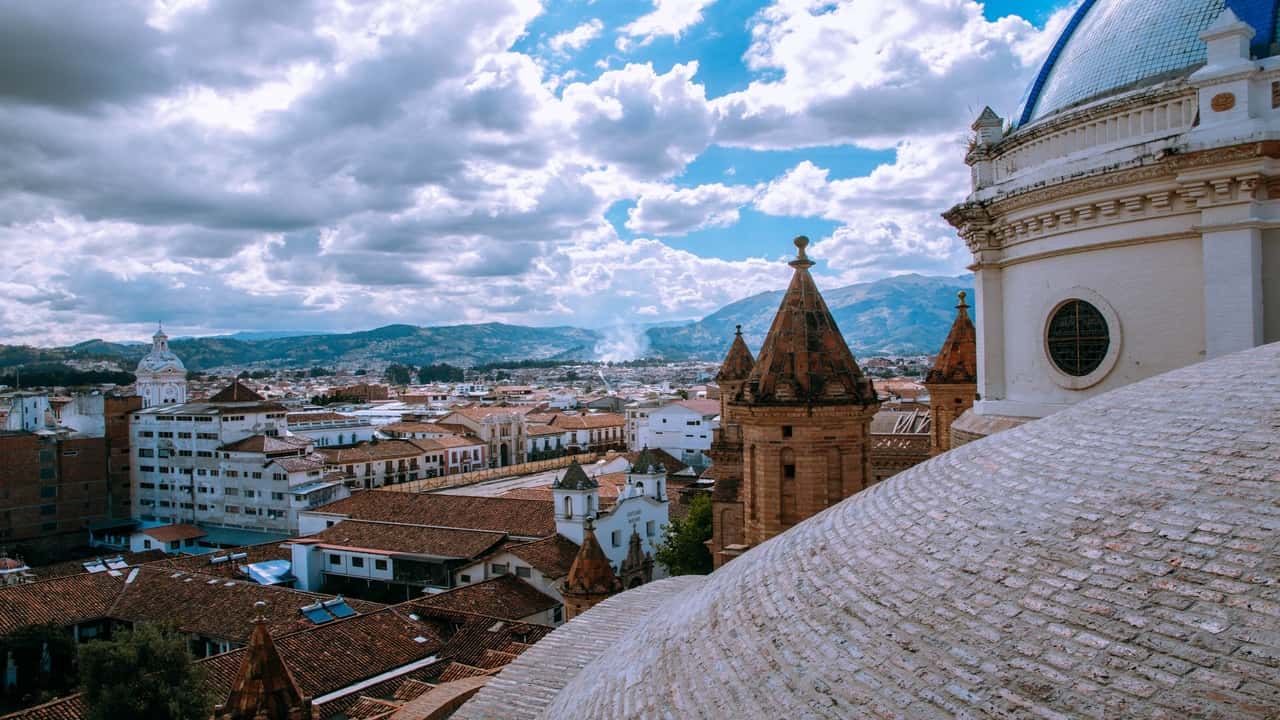 A panoramic view of a city with historical architecture, surrounded by mountains under a cloudy sky