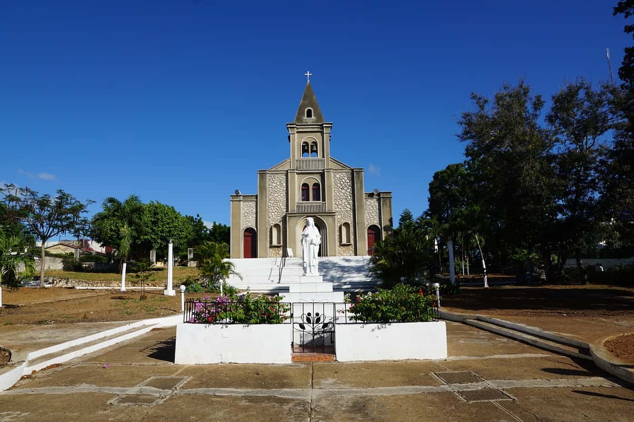 A church with a central statue in front, surrounded by greenery under a clear blue sky