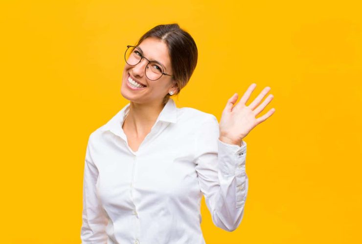 The image shows a smiling person waving, dressed in a white button-up shirt with glasses, standing against a bright yellow background.