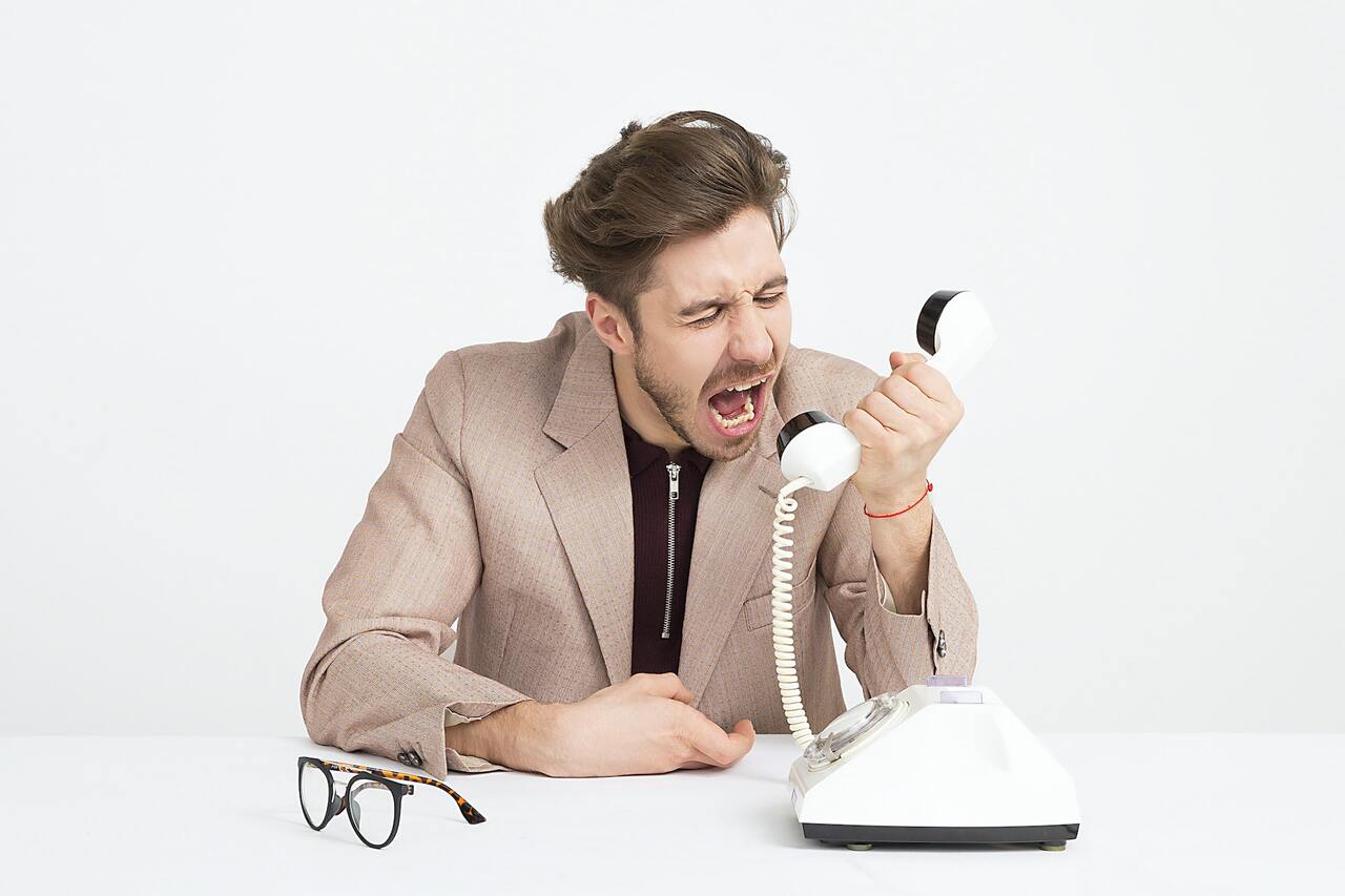 A man in a beige suit is angrily yelling into a white rotary telephone