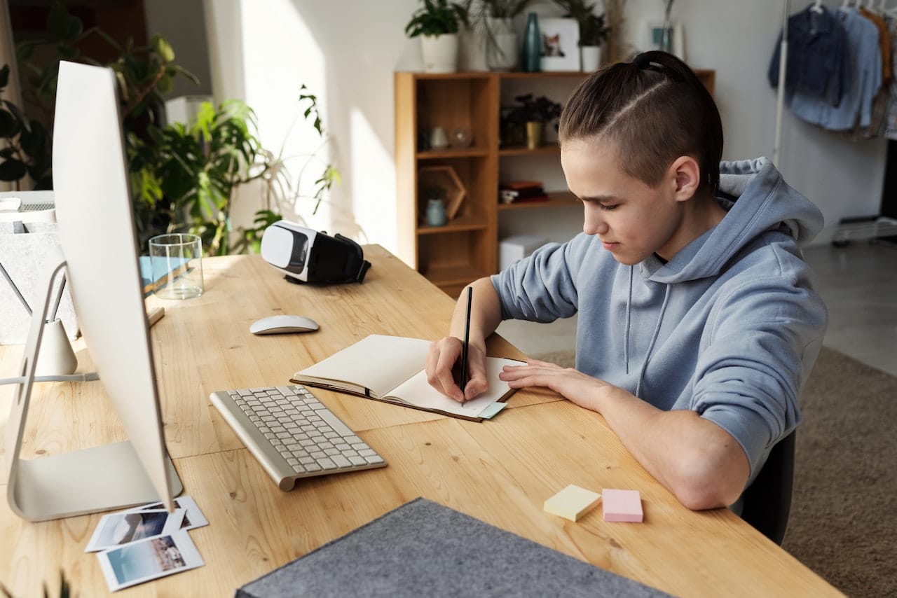 Person writing notes at a desk with a computer