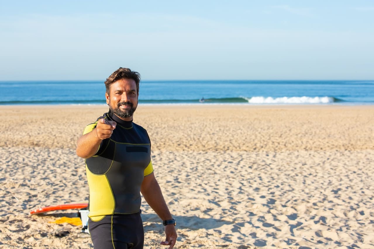 Man in wetsuit standing on beach pointing at the camera