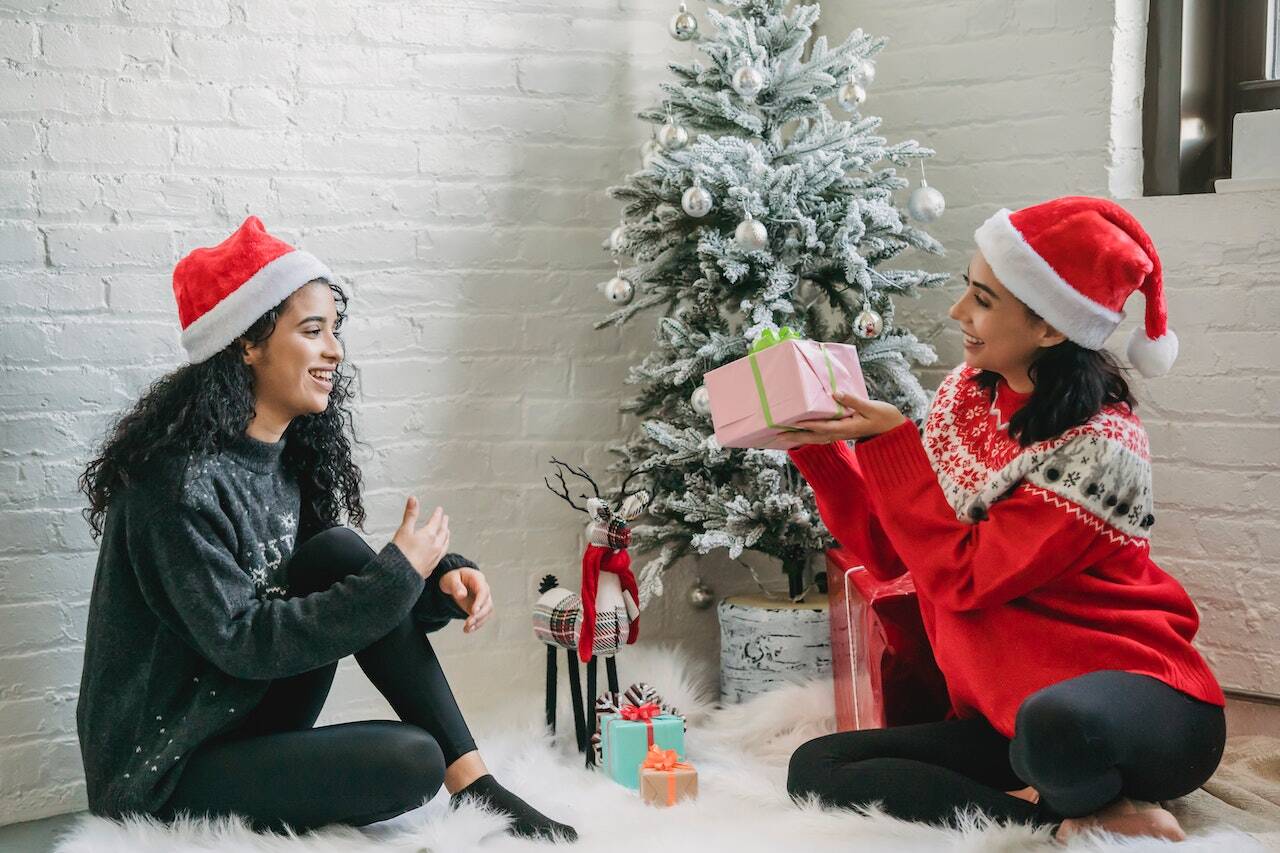 Two women wearing Santa hats exchange a gift near a decorated Christmas tree.