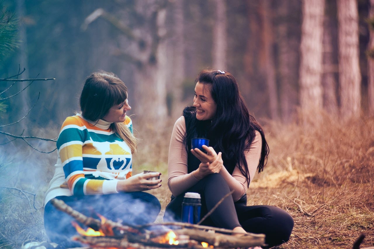 Two women sitting by a campfire with drinks