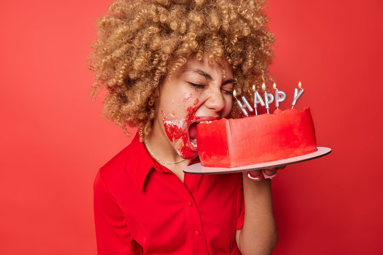 Person with curly hair biting into a red birthday cake