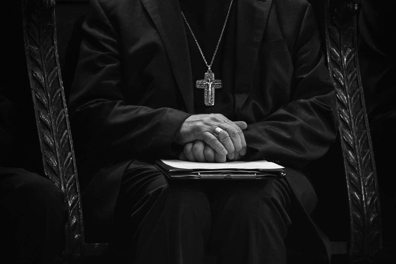 Clergyman seated with hands folded and wearing a large cross