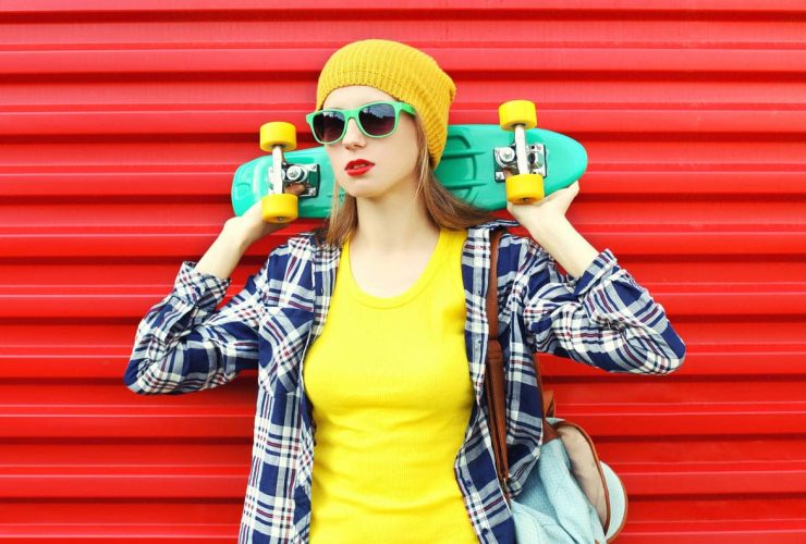 Young woman in sunglasses holding a skateboard against a red background.