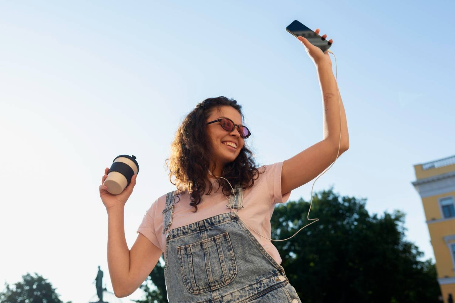 Person holding phone and cup while smiling outdoors.