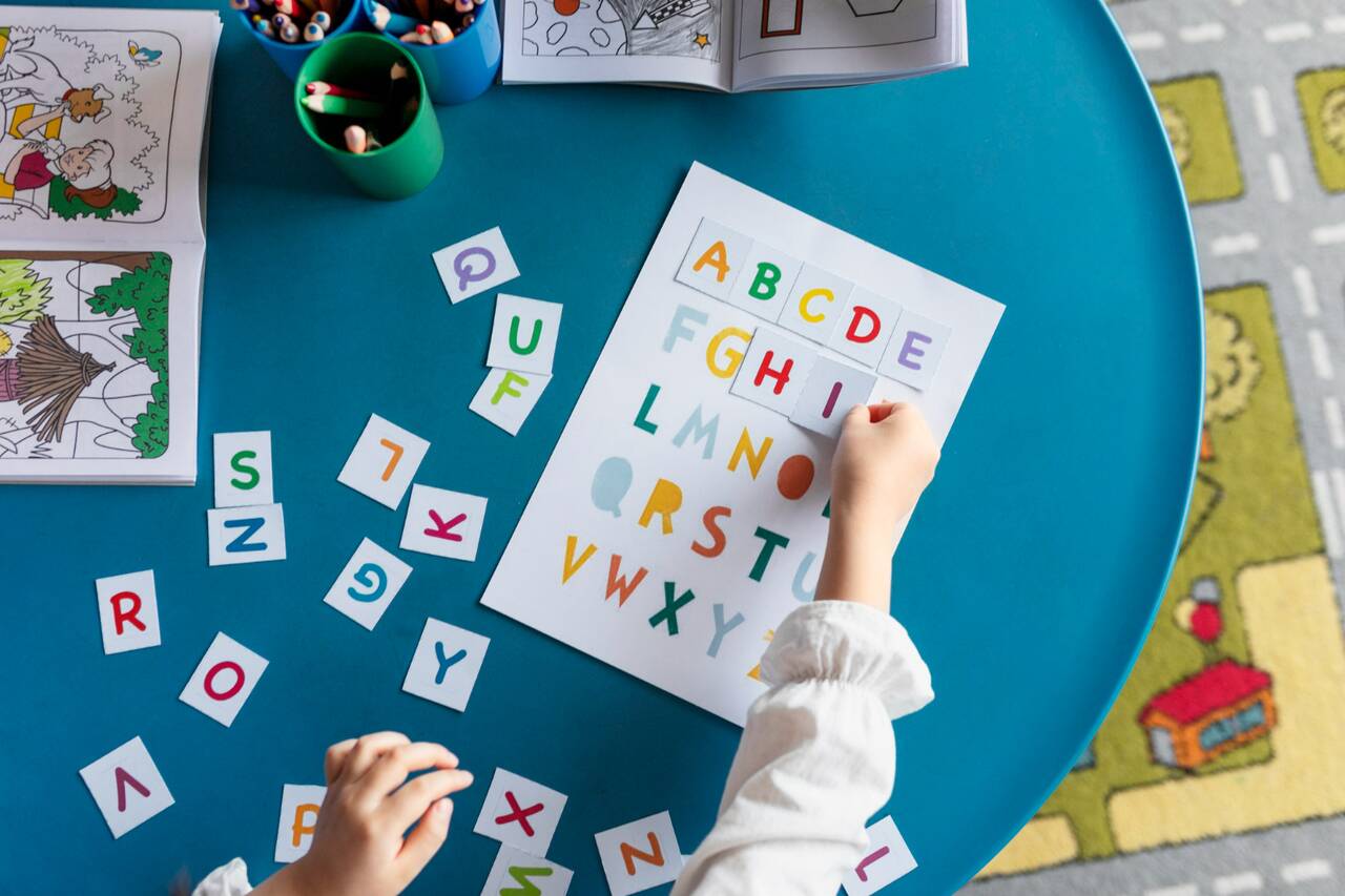 A child arranging colorful alphabet cards on a worksheet on a blue table.