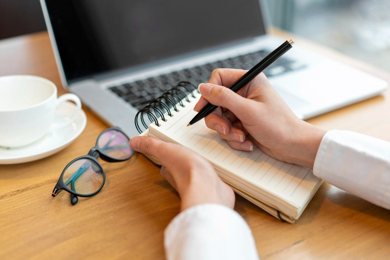 Person writing in a notebook near a laptop, glasses, and a cup