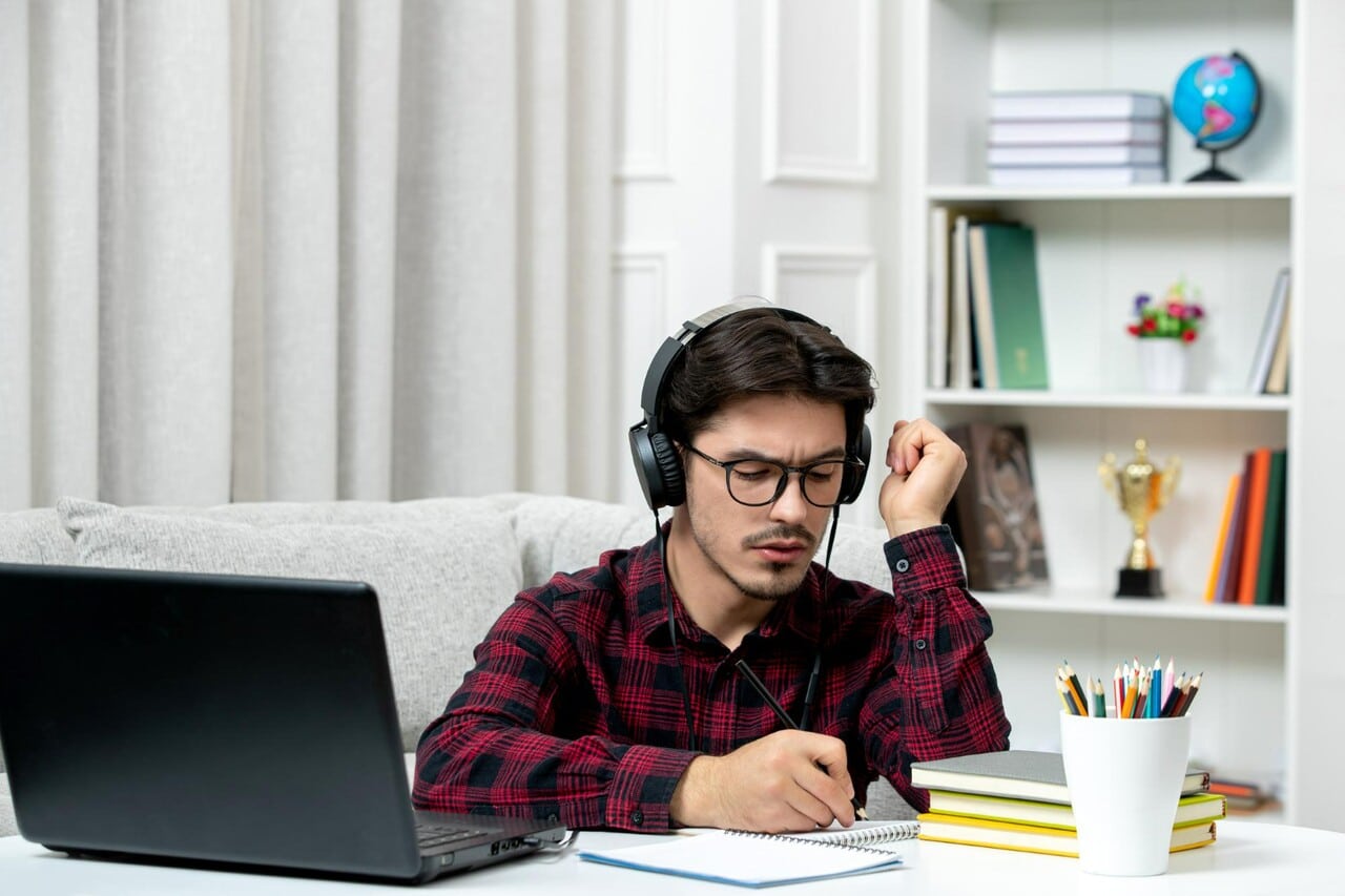 Focused student writing notes while wearing headphones in front of a laptop