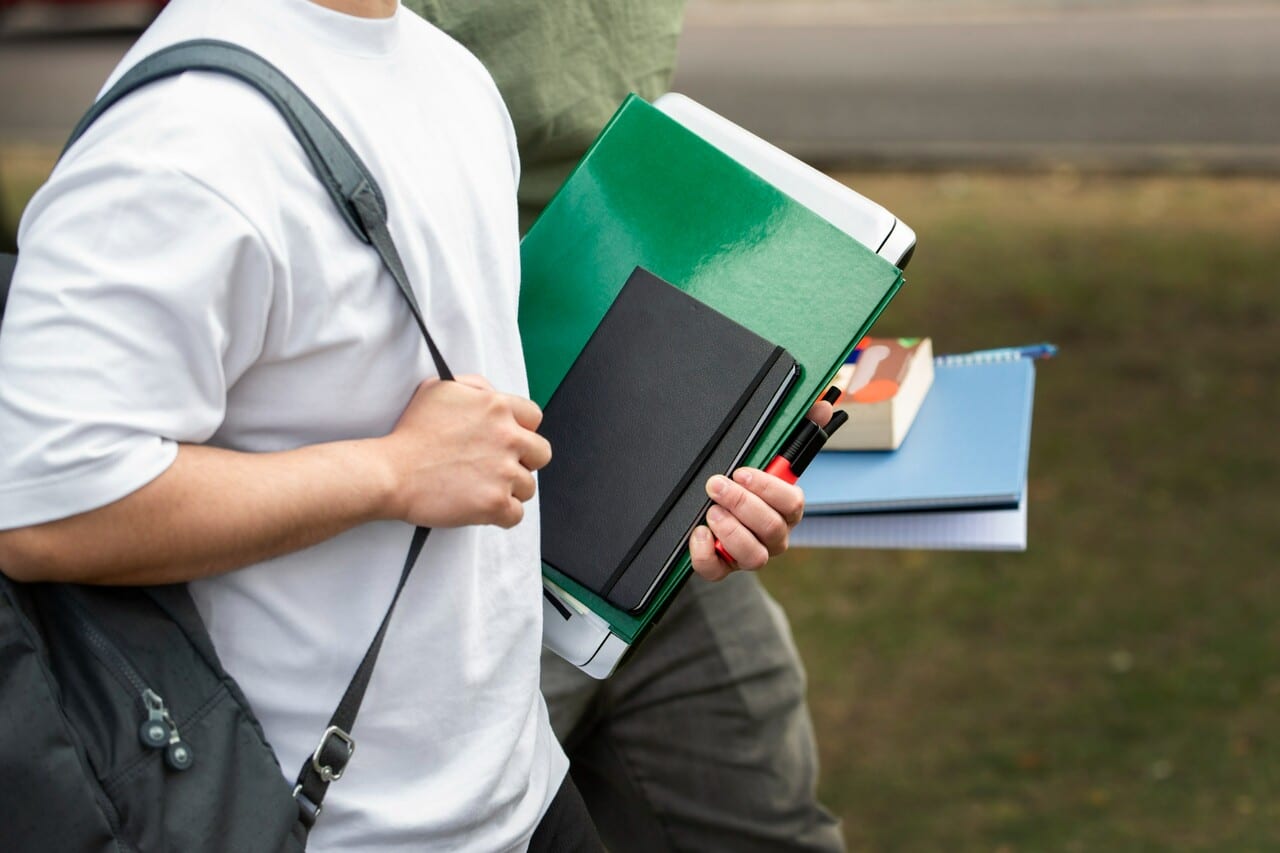 Two students carrying notebooks and binders while walking, emphasizing a close-up of their books and backpacks.