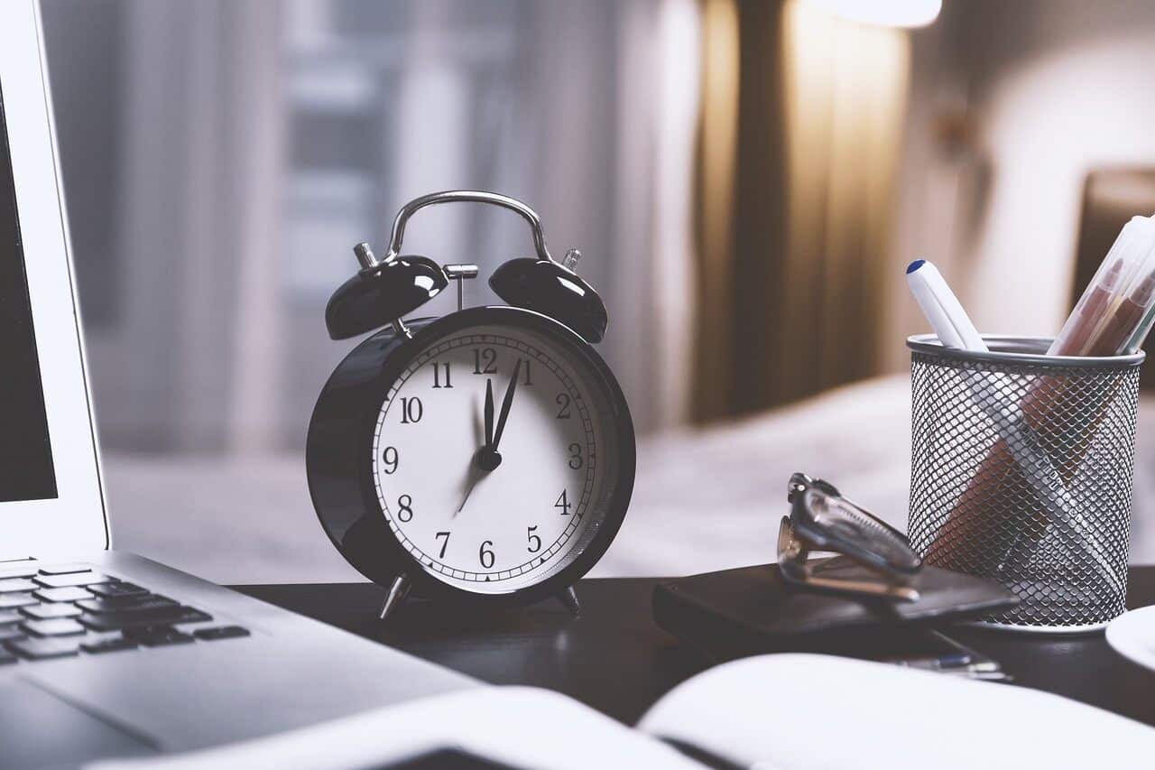 Black alarm clock on desk beside a laptop and pen holder.