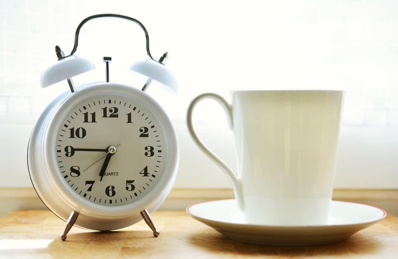 White alarm clock beside a white coffee cup on a wooden table.