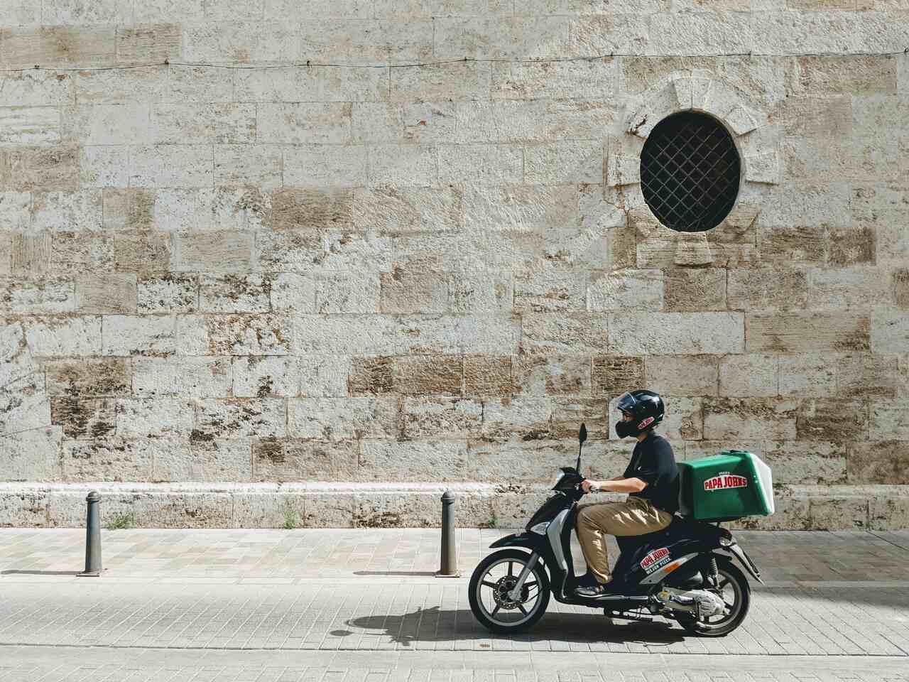 Delivery driver riding a scooter with a Papa John’s box on the back, passing a rustic stone wall