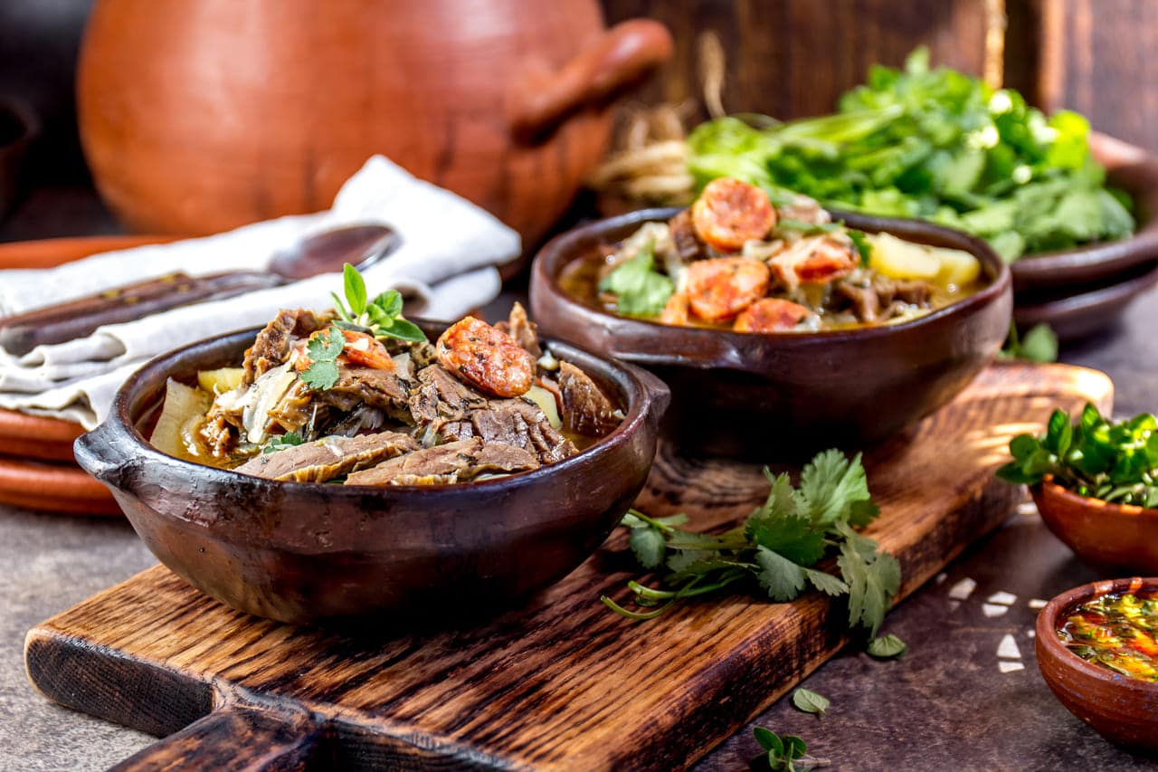 Rustic wooden table with two bowls of hearty stew, garnished with herbs, accompanied by fresh greens.