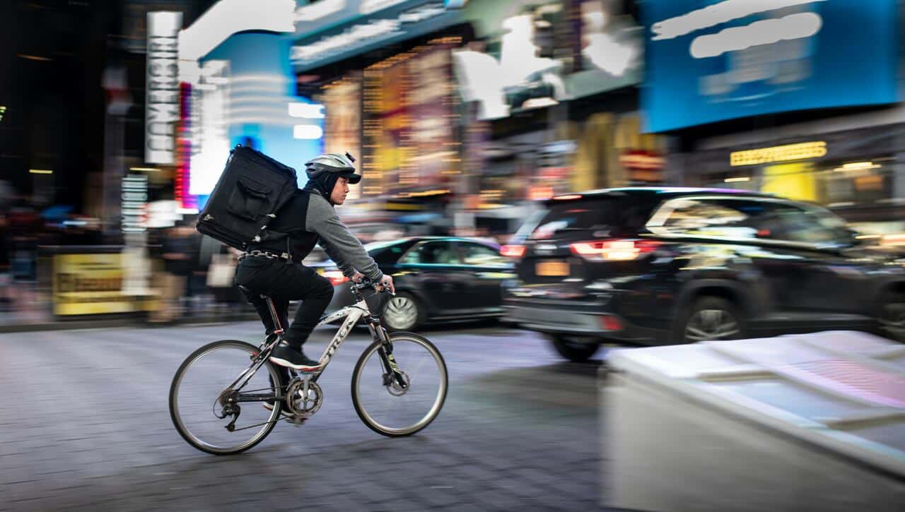 Food delivery cyclist navigating a busy city street at night, with bright lights and moving traffic around.