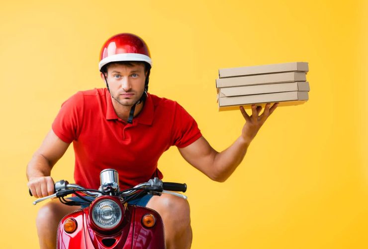 Male delivery driver in a red helmet holding pizza boxes while sitting on a scooter, against yellow background.