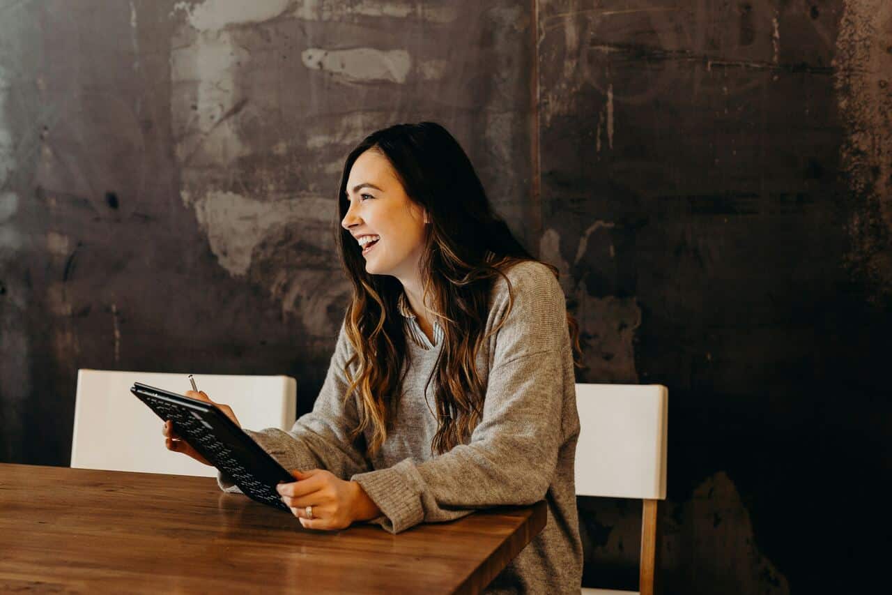 A woman smiles while holding a tablet, sitting at a wooden table, enjoying conversation indoors