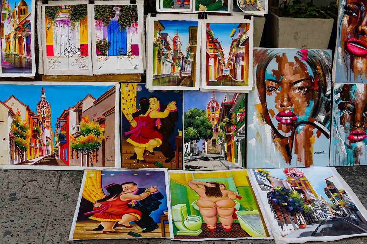 Colorful display of various paintings, including vibrant street scenes, dancing figures, and abstract portraits, arranged on the ground and against a wall