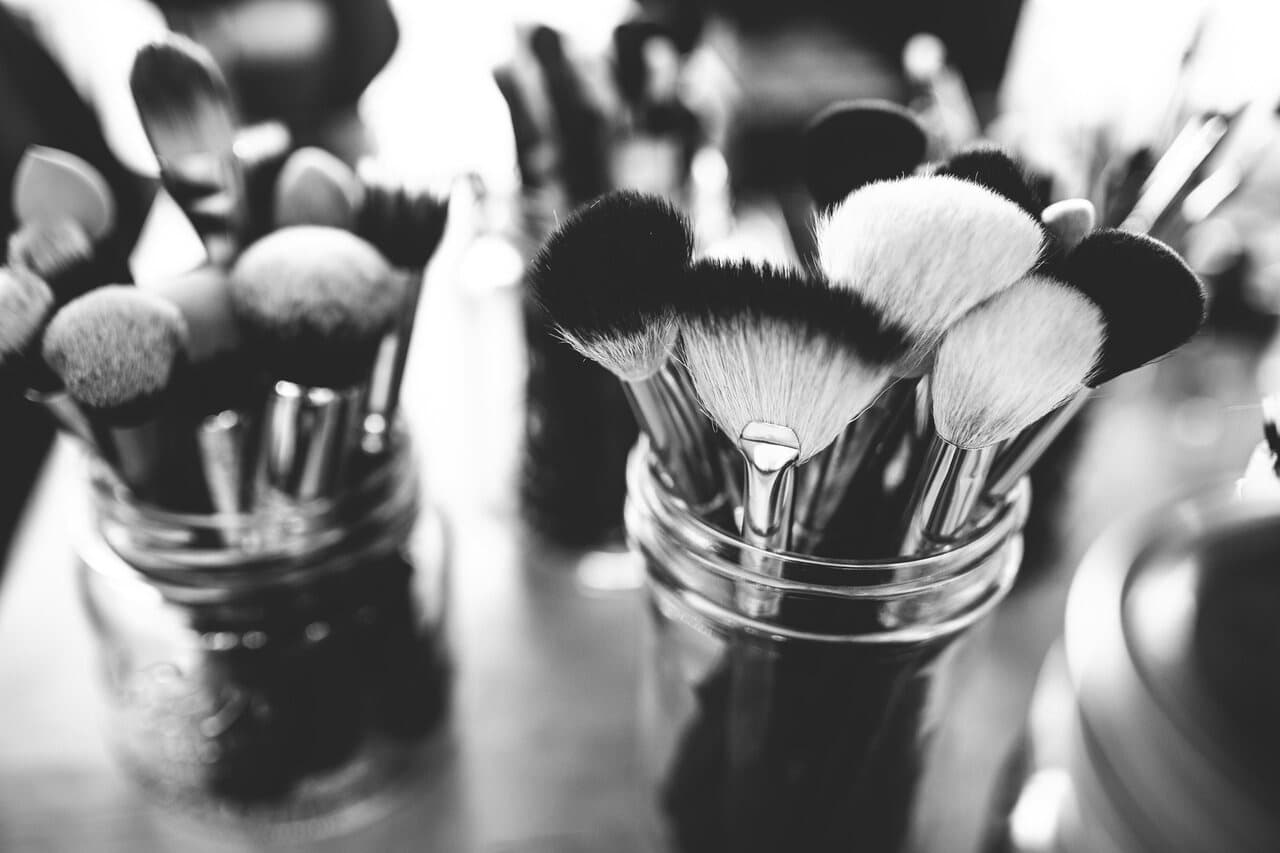 Close-up of various makeup brushes in glass jars, arranged on a tabletop in black and white