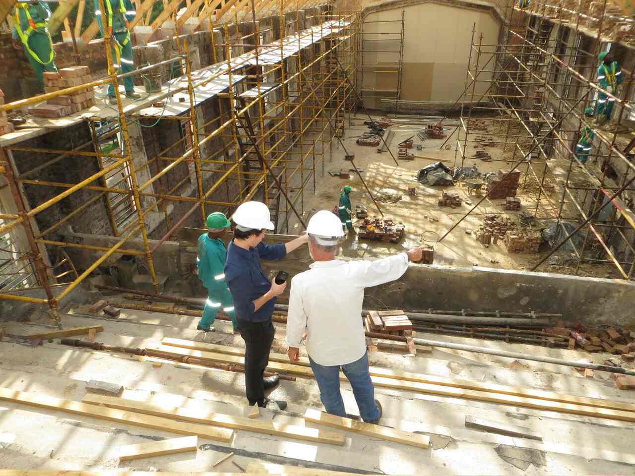 Two construction supervisors in white hard hats discussing a building site, with scaffolding, workers, and construction materials like bricks and wooden planks visible in the background