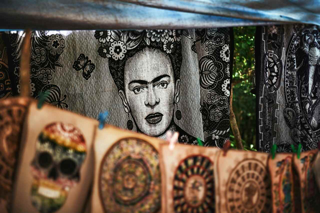 Black and white tapestry featuring a portrait of Frida Kahlo with floral patterns, displayed behind colorful artwork, including skull and circular designs in the foreground