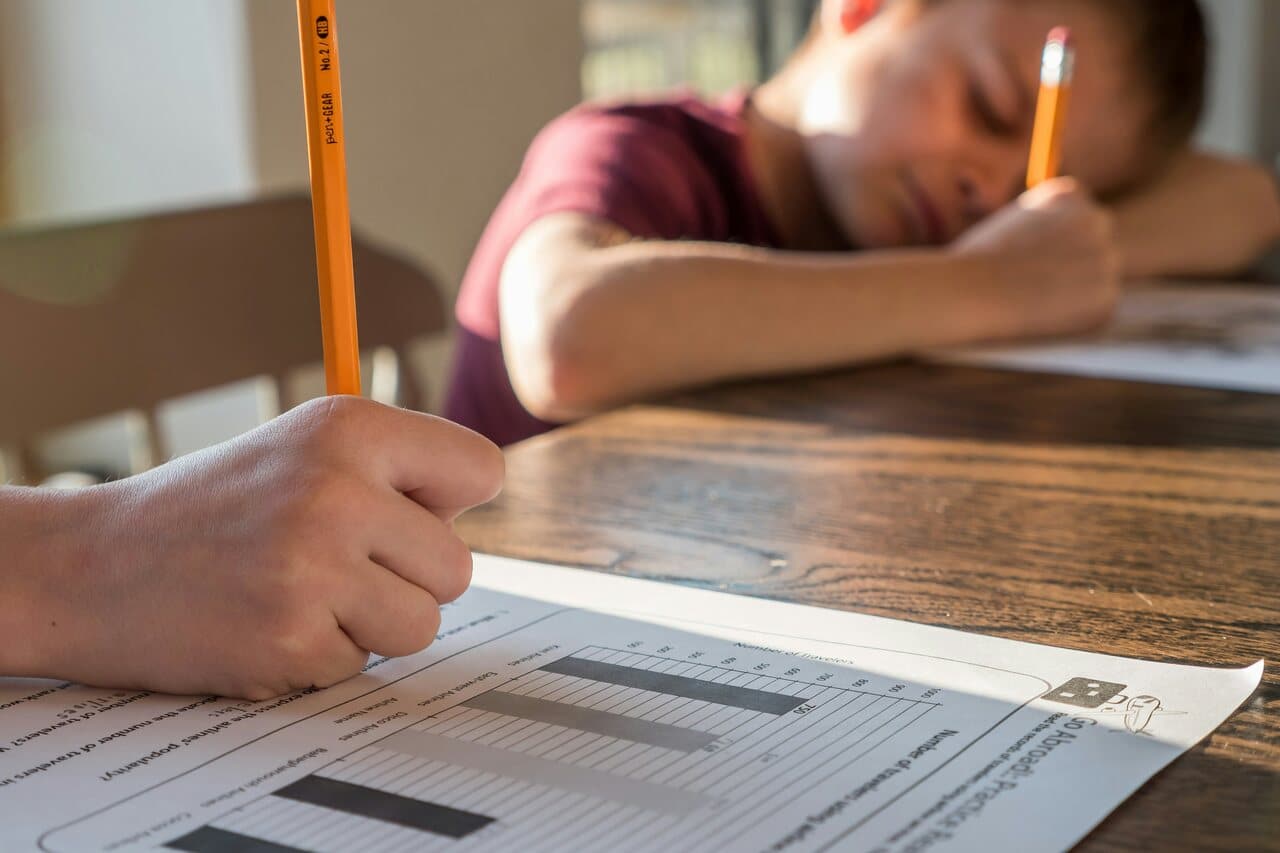 Two children working on worksheets at a table, with one focusing on writing with a pencil while the other rests their head on the table, appearing tired.