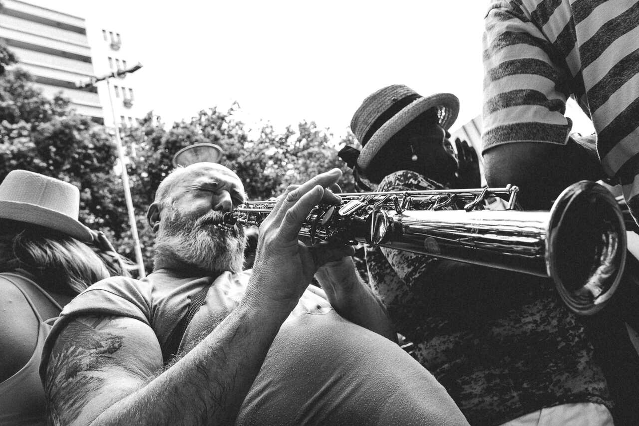 Bearded musician intensely playing a saxophone outdoors during a street performance, surrounded by people.