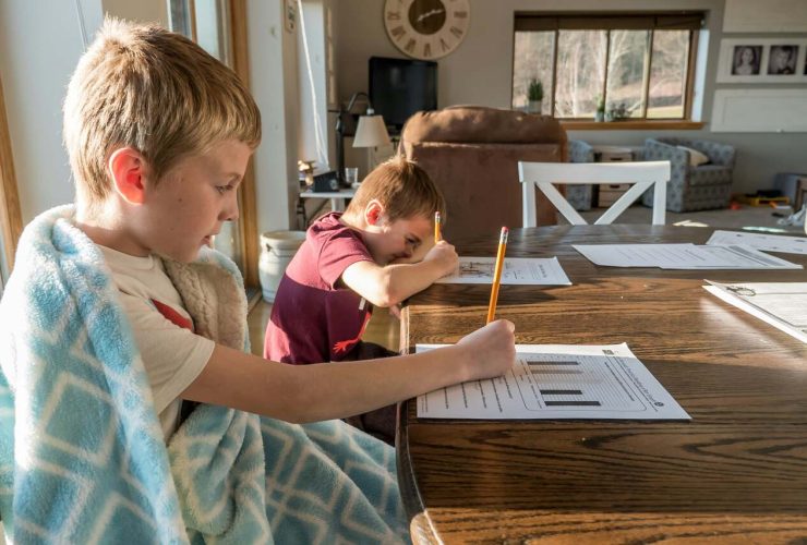 Two boys working on worksheets at a wooden table, one wrapped in a blue blanket and the other resting his head on his arm while writing with a pencil, in a well-lit living room