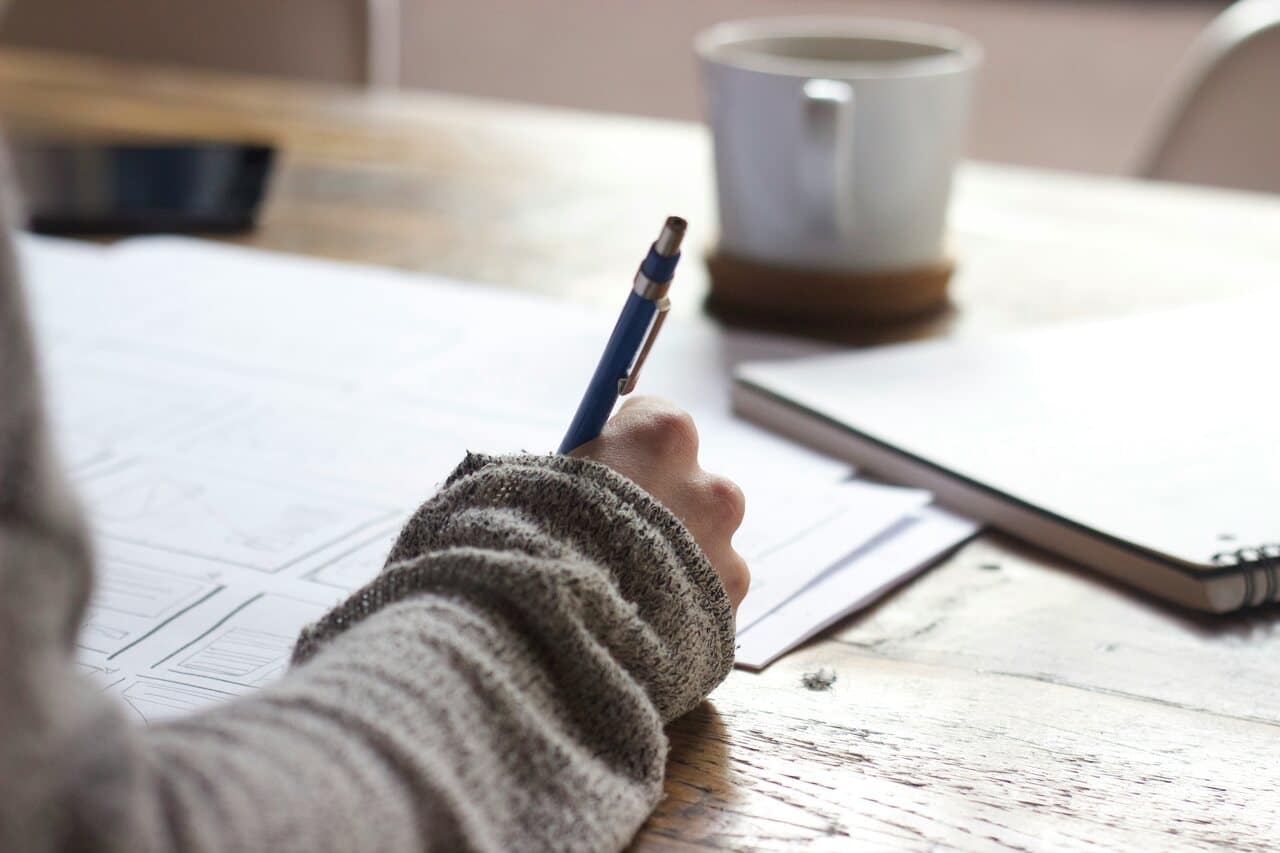 A person writes with a pen on paper at a desk, with a coffee cup nearby.