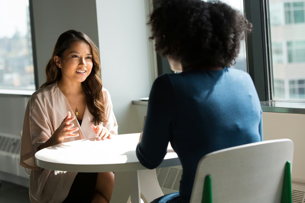 Two women having a discussion at a round table in a bright office space, with one facing the camera and the other with her back turned.