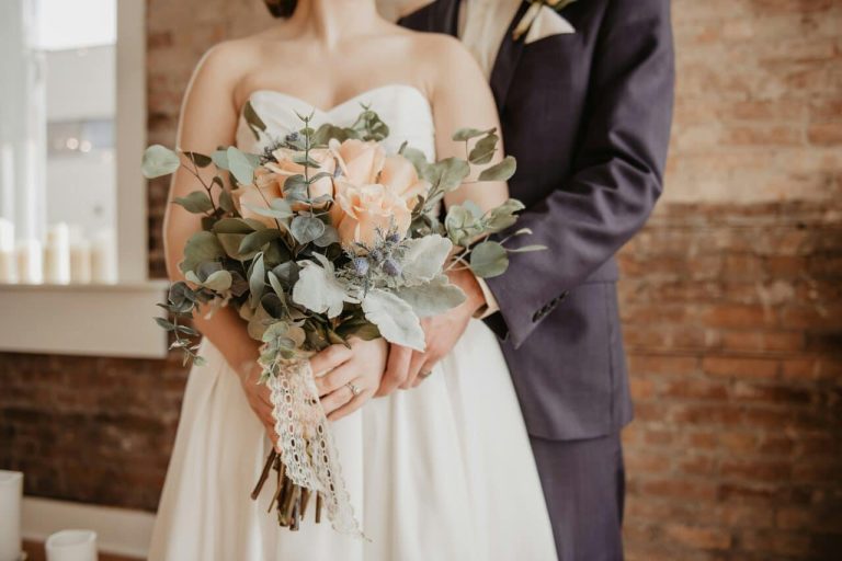 Bride holding a bouquet of peach roses and greenery, while groom stands behind her, embracing gently.