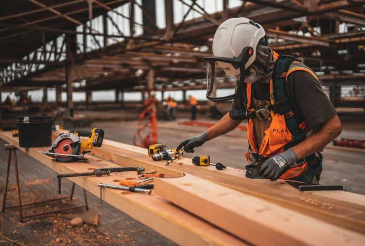 Construction worker wearing safety gear, including a hard hat and face shield, using power tools to work on wooden beams at an industrial construction site, with tools like drills and saws scattered on the table.