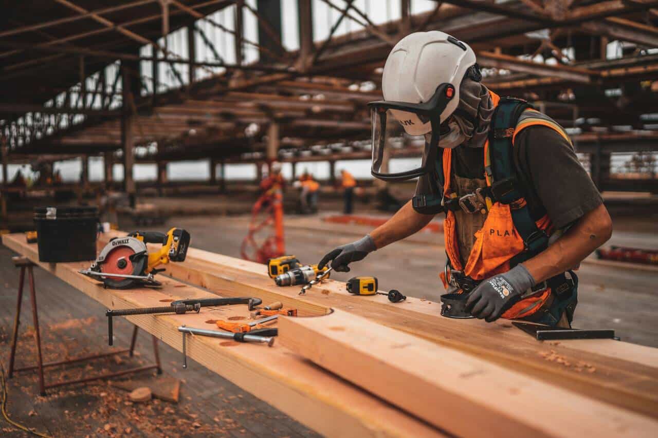 Construction worker wearing safety gear, including a hard hat and face shield, using power tools to work on wooden beams at an industrial construction site, with tools like drills and saws scattered on the table.