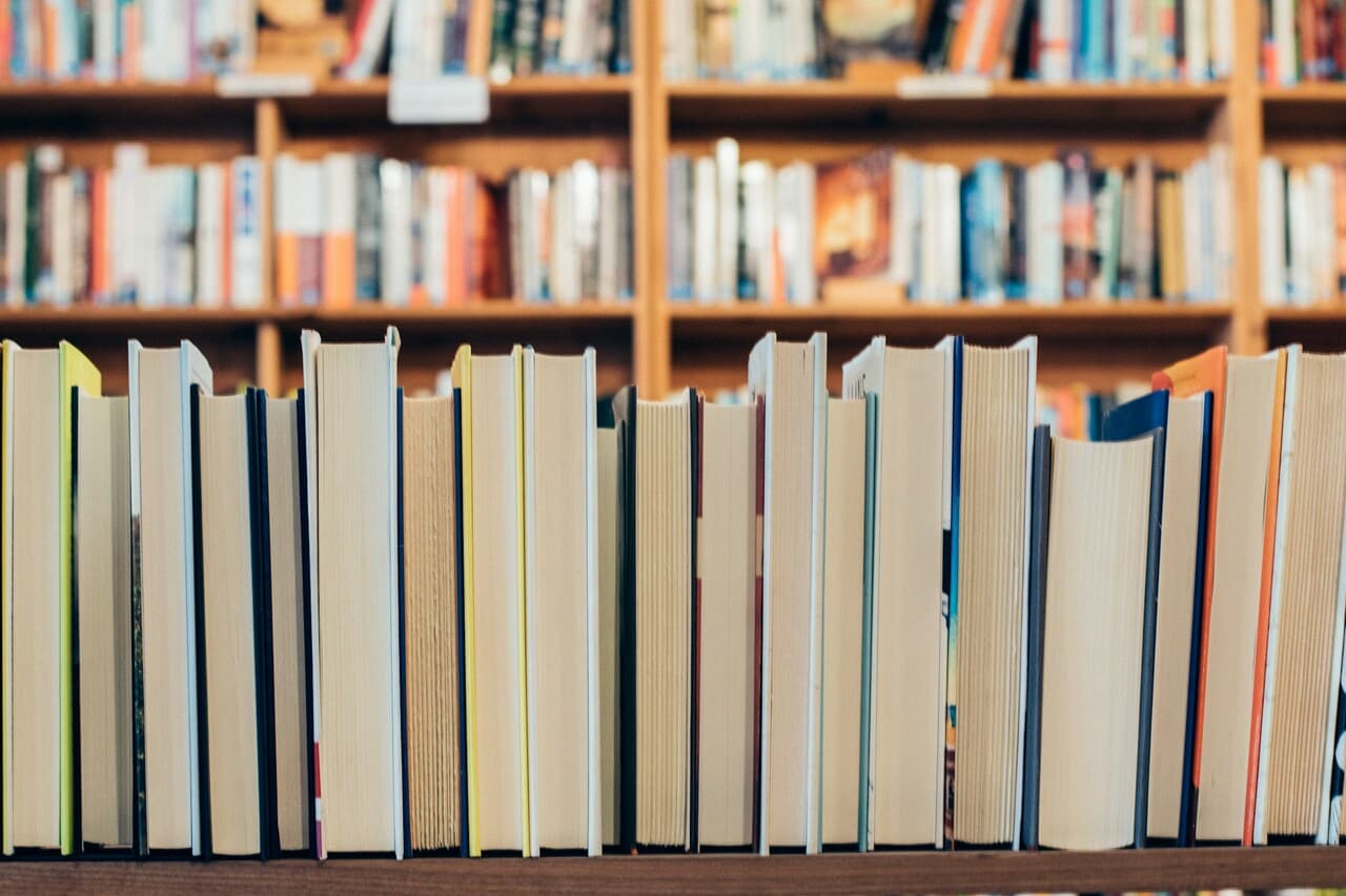 Books neatly arranged on a shelf with their spines facing down, with a blurred library background of additional books on shelves