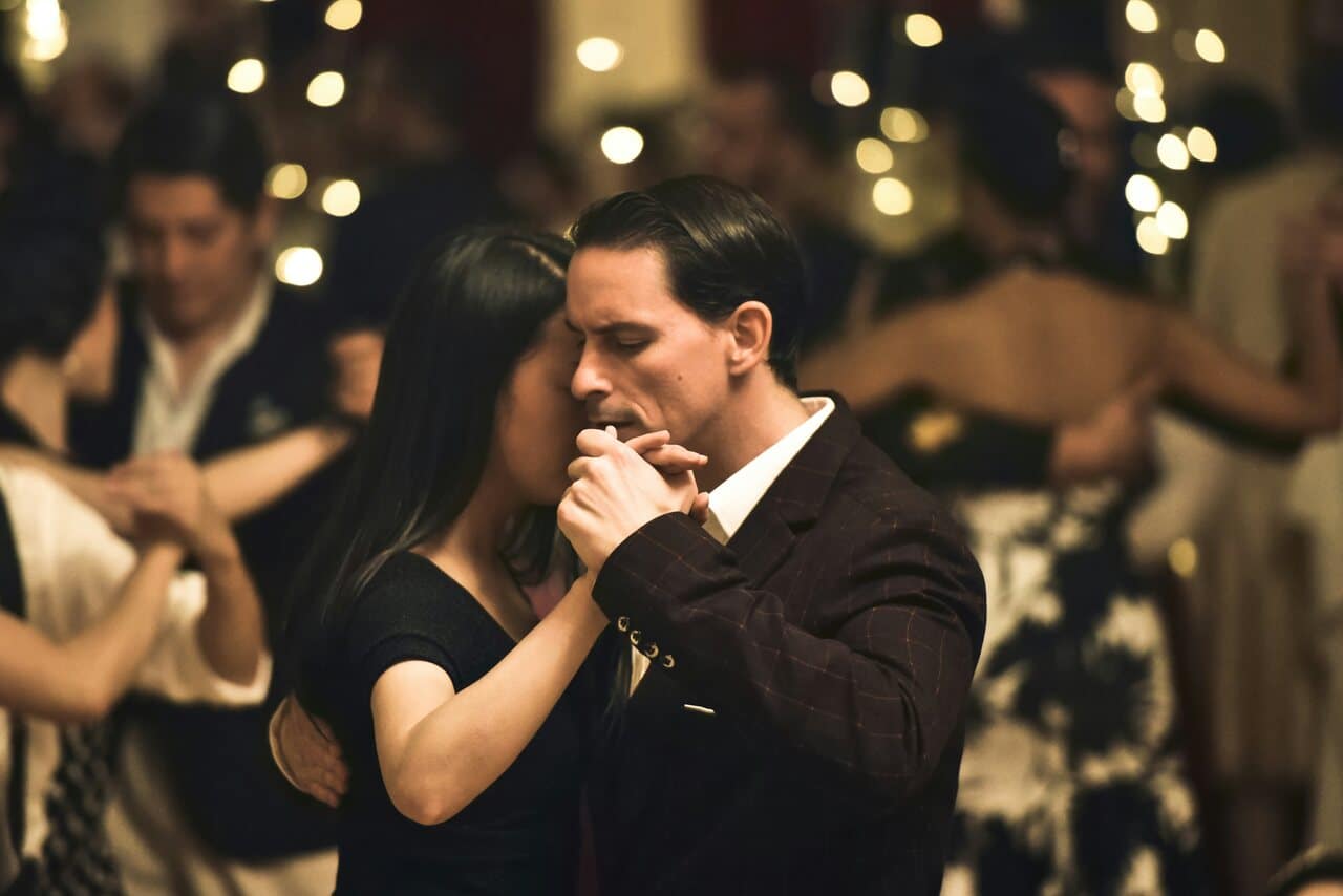 Couple dancing closely at a formal event, surrounded by other dancers under soft string lights.