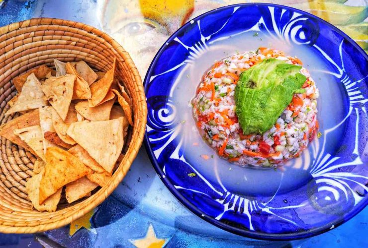 Mexican ceviche topped with avocado, served on a blue decorative plate, accompanied by a basket of tortilla chips on a colorful table.