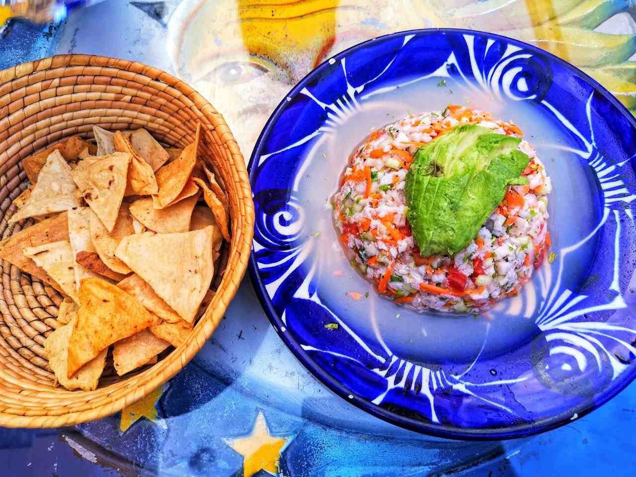 Mexican ceviche topped with avocado, served on a blue decorative plate, accompanied by a basket of tortilla chips on a colorful table.